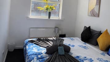 A bedroom with a bed, a window with flowers, a radiator, and a painting on the wall.