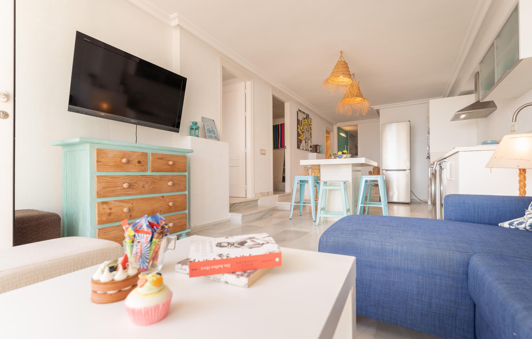 A modern living room with a flat-screen TV, a teal chest of drawers, a blue sofa, and a kitchen area in the background.