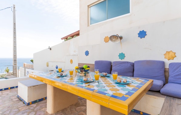 A dining area with a tiled table, purple cushions, and a view of the sea.