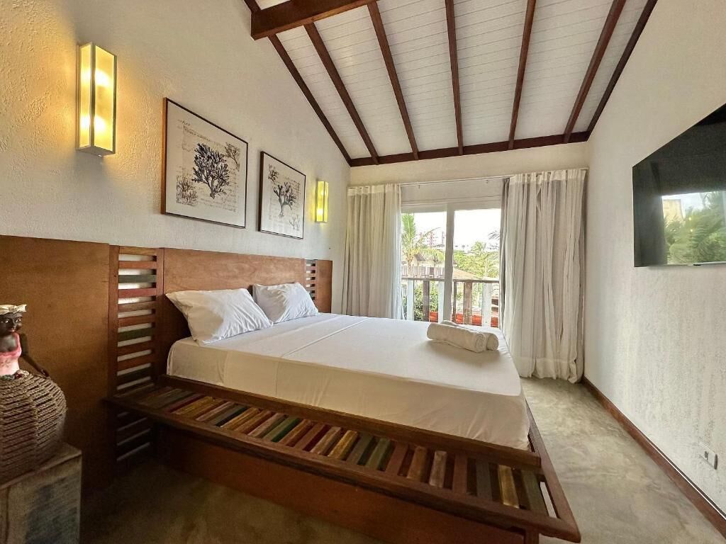 Un dormitorio con cama de madera, ropa de cama blanca, una mesita de noche, luces fijadas a la pared e impresiones botánicas enmarcadas.