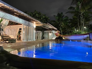A swimming pool with a covered area and lounge chairs, surrounded by palm trees.