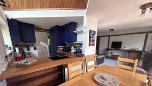 Een compacte keuken met houten kastjes, een eettafel met stoelen en een televisie in de woonkamer.