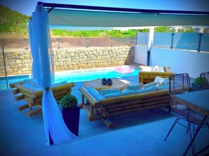 Ein überdachter Außenbereich mit Pool, Holzmöbeln und einer Wand aus Stein im Hintergrund.