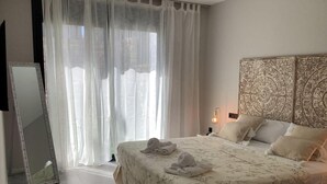 Quarto com cama, mesa de cabeceira, luminária, espelho e janela com cortinas.