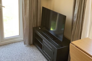 A room with a television on a black stand, a wooden cabinet, and a window with curtains. - Yeovil 1-Bed Annex Sleeps 2 with WiFi (Yeovil)