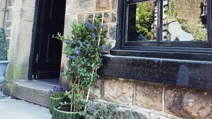 En bygning i sten med sortmalede vinduesrammer, en dør og en lille have med lilla blomster.