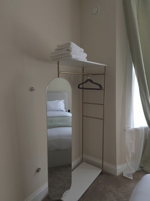 Een compacte kamer met een bed, een kleine inloopkast met een spiegel, en een raam met dunne gordijnen.