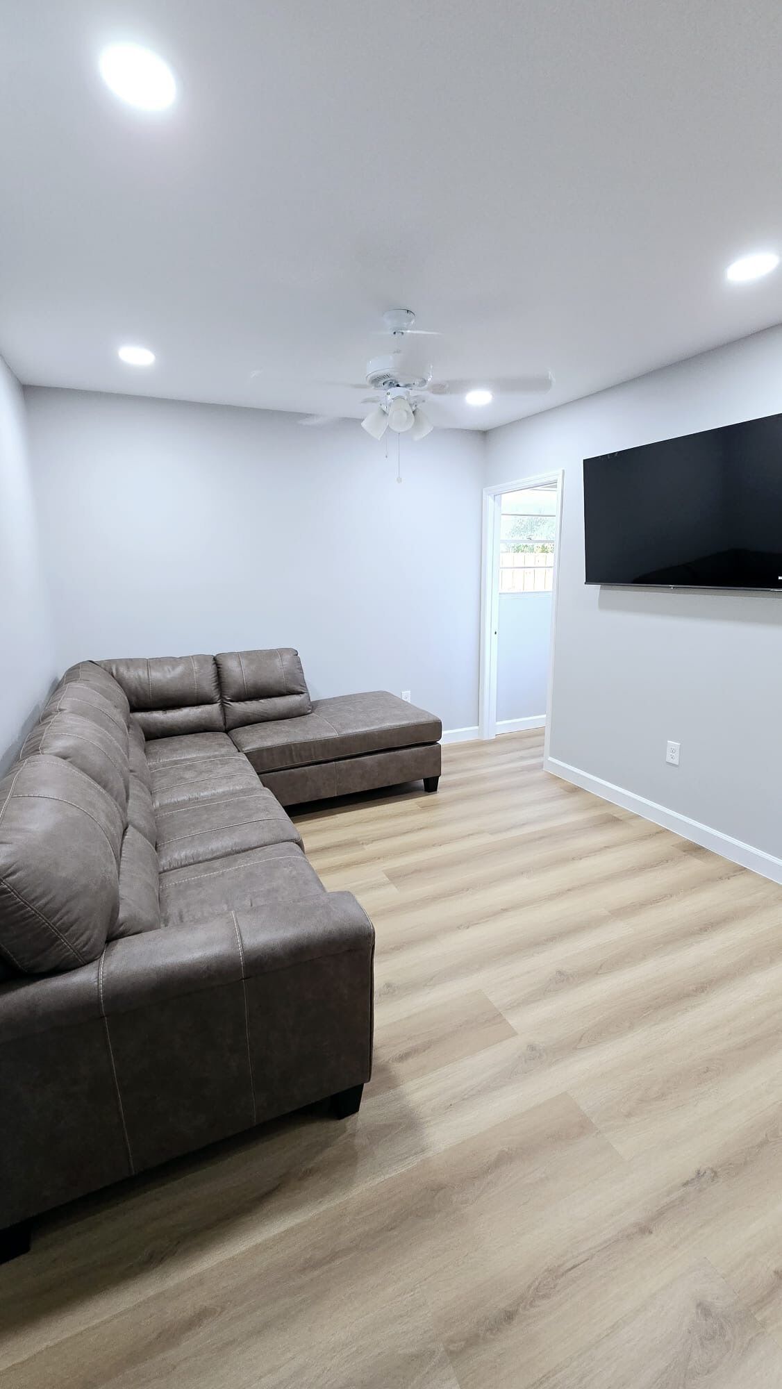 A modern living room with a sectional sofa, a ceiling fan, recessed lighting, and a flat-screen TV mounted on the wall.