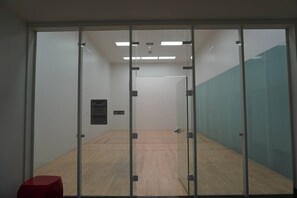 Een squashbaan met glazen wanden, een houten vloer en een rode stoel.