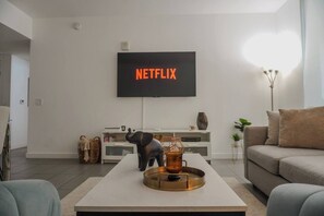 Ein modernes Wohnzimmer mit einem an der Wand befestigten Flachbildfernseher, der 'NETFLIX' in orangefarbenen Buchstaben anzeigt, einem Couchtisch mit einem dekorativen Gegenstand und einem Sofa.