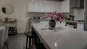 A modern kitchen with white cabinets, a central island with a vase of flowers, and a dining area with a chair and a small table. - Luxurious 1BR condo in prime Location (Miami)