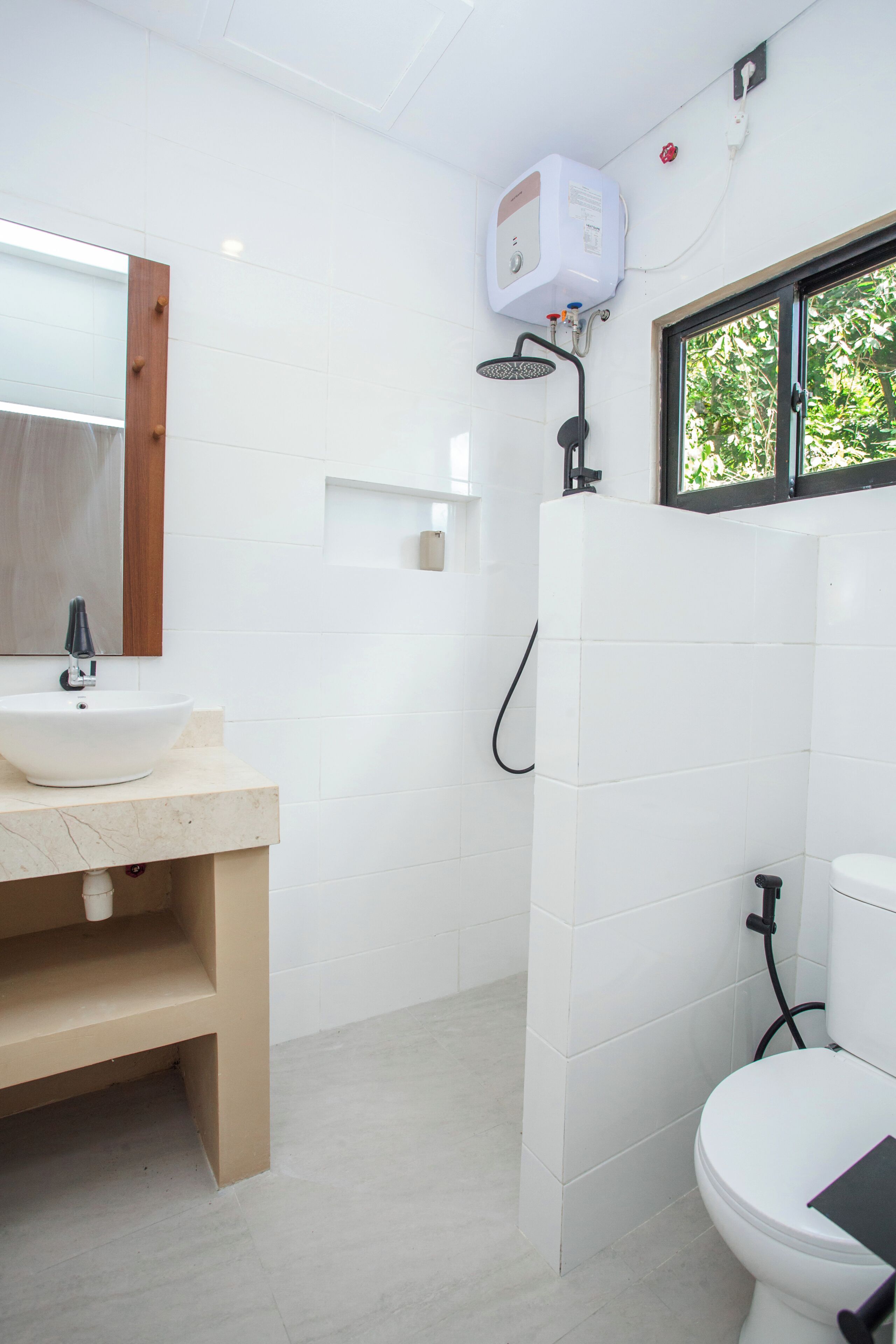 Ein Badezimmer mit Waschbecken, Spiegel und Toilette, sowie einer Dusche mit Duschschlauch und einem Fenster mit Blick auf Grünflächen.