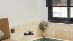 Una habitación con una cama, un ventanal pequeño, un lavamanos y un florero con flores.