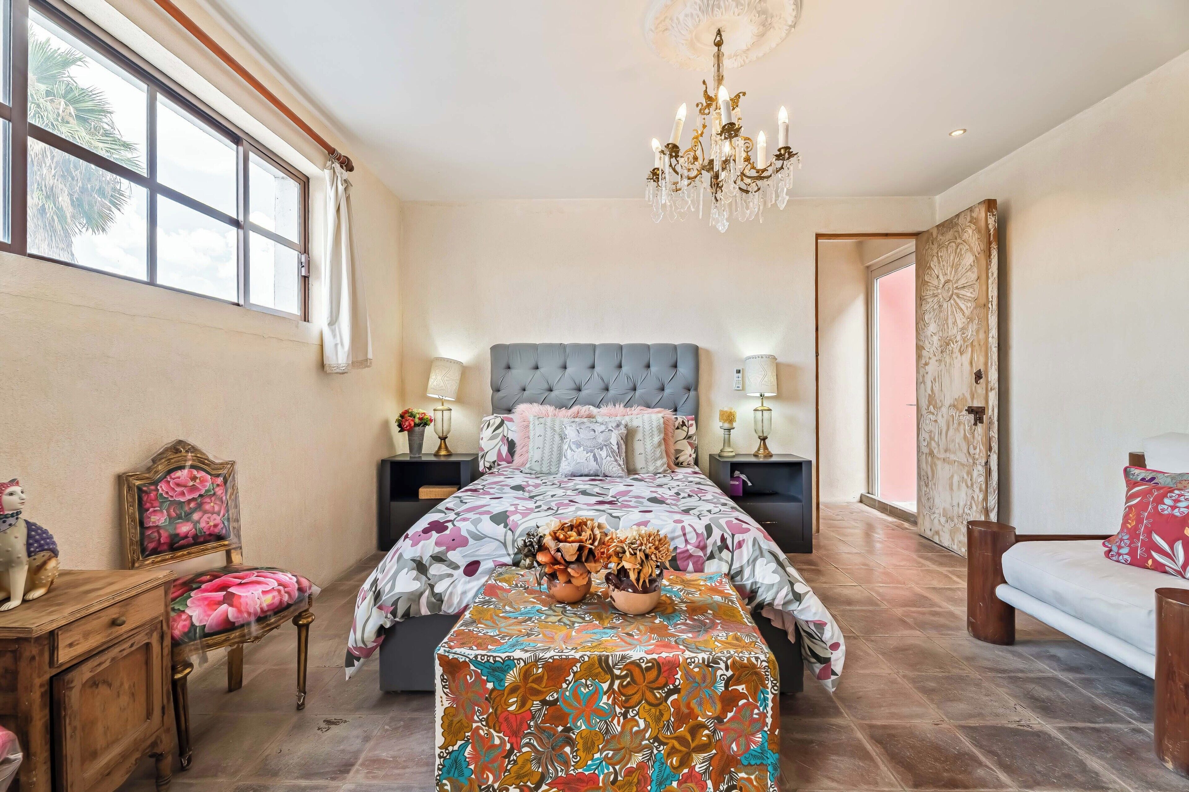 A bedroom with a large bed, a chandelier, a window with curtains, a wooden dresser, and a chair.