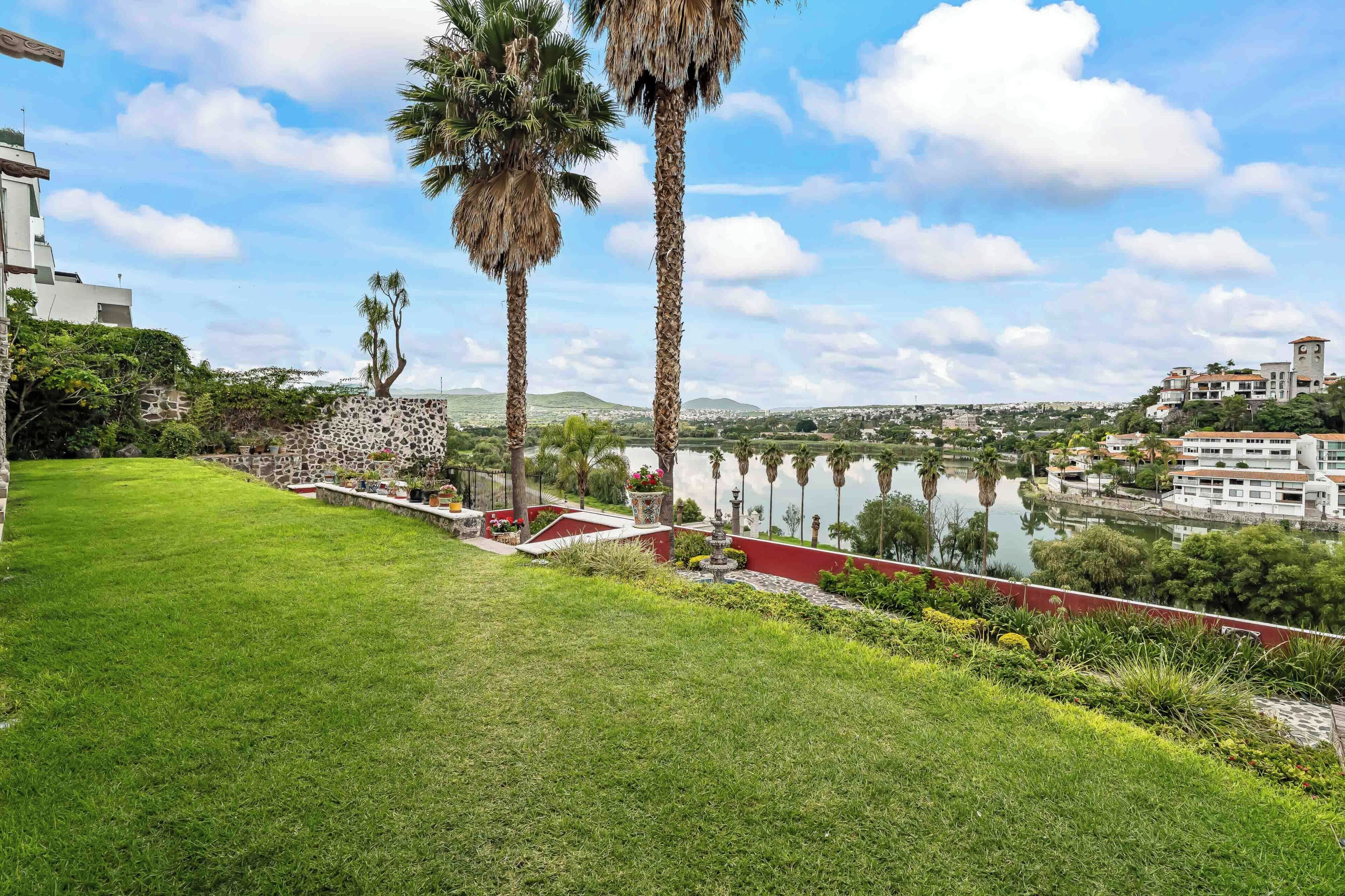 A well-maintained lawn, palm trees, a stone wall, and a view of a lake with buildings in the background.