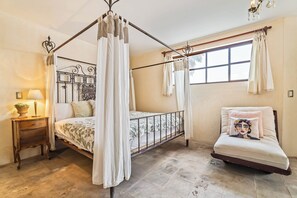 A bedroom with a four-poster bed, a bedside table with a lamp, a chair with a cushion, and a window with curtains.