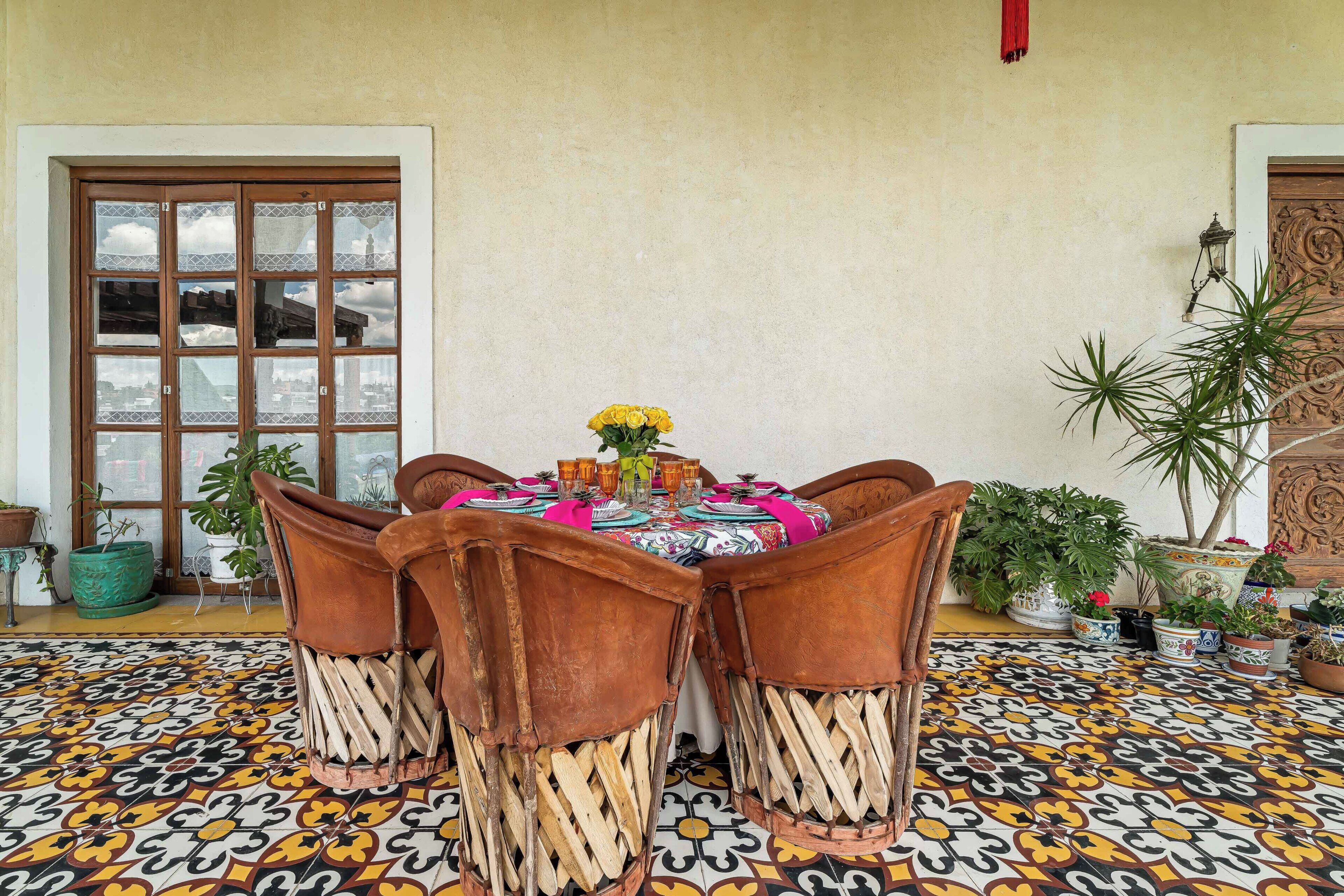 A dining area with a table set for four, surrounded by chairs with woven bases, a patterned floor, and potted plants.