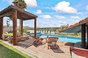 A poolside area with wooden loungers, a pergola, and a view of a lake and distant buildings.