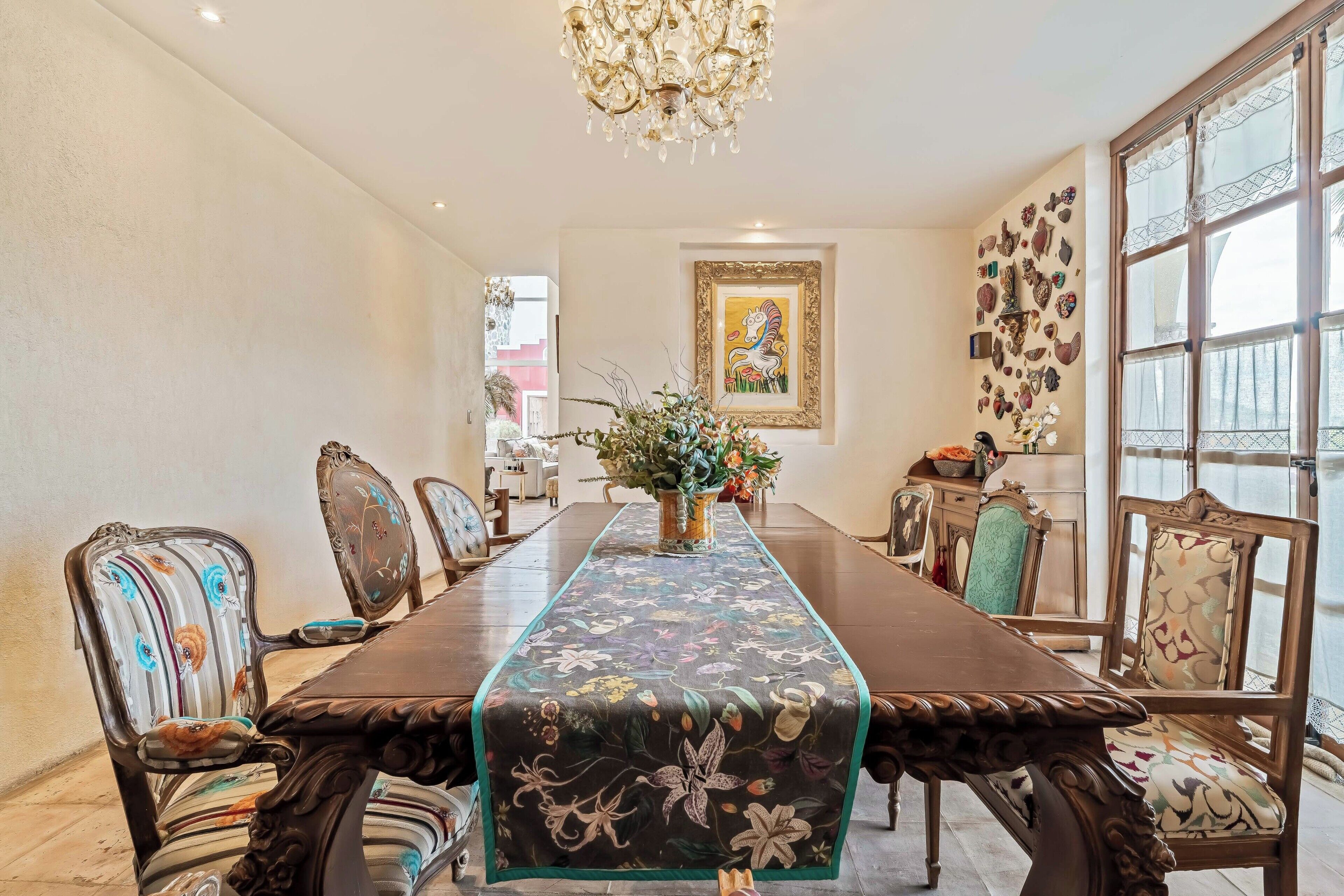 Une salle à manger avec une longue table, des chaises élégantes, un lustre et un mur orné d’objets décoratifs.