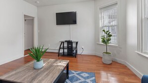 A room with a wooden floor, a dining table with a blue rug, a black TV stand, and a potted plant near a window. - The Red Door Retreat - Walking Distance to Downtown Hartford (Hartford)