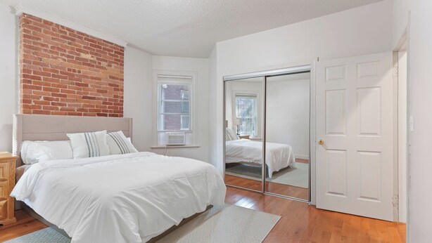 A bedroom with a bed, a wooden nightstand, a brick wall, a window, a sliding glass door, and a white door. - The Red Door Retreat - Walking Distance to Downtown Hartford (Hartford)