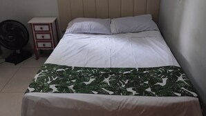 Ein Einzelbett mit Kopfteil, ein Nachttisch mit Schubladen, ein Standventilator und ein Fenster mit Holzjalousien.