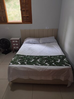 A single bed with a headboard, a bedside table with drawers, a standing fan, and a window with wooden shutters. - Beautiful country house in the Porta do Sol gated community. (Mairinque)