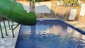 Ein Swimmingpool mit einer grünen Rutsche, Steinmauern und einer gefliesten Liegefläche.