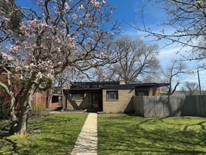 A small house with a front yard, a wooden fence, and a tree with pink blossoms. - Beautiful Mid-Century Oasis in South Shore. (Chicago)