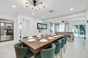 A dining area with a long table set for six, green upholstered chairs, a ceiling fan, and a view of the outdoors. - Spacious 5BR Heated Pool Villa · minutes to Beach (Hollywood)