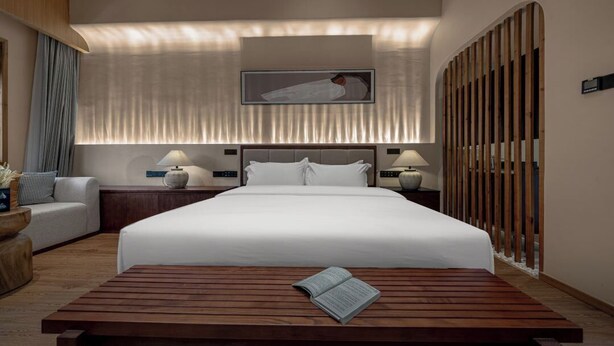 Habitación de hotel con una cama grande, lámparas de noche, una mesita de madera con un libro, un sofá y un panel decorativo en la pared.