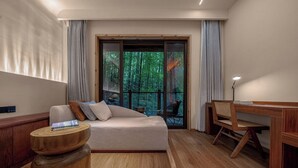 Una habitación moderna con sofá, mesita de madera y escritorio con silla. Hay un ventanal con vistas a árboles.