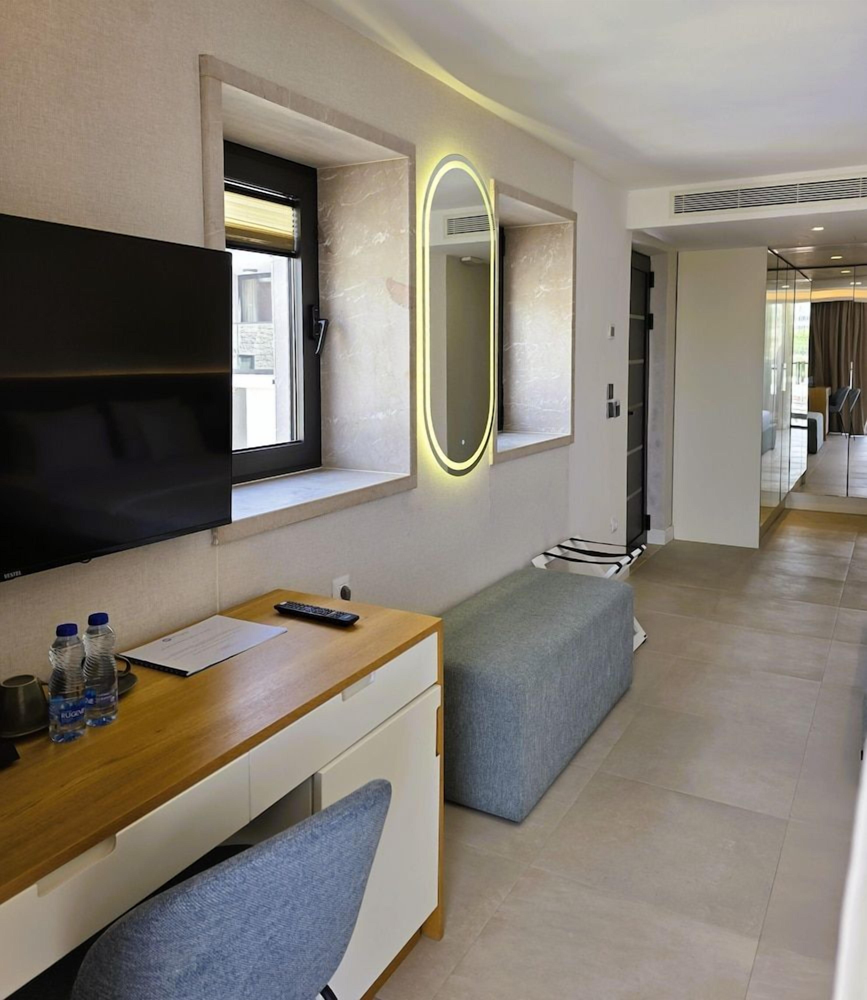 A modern hotel room with a flat-screen TV, a desk with a notebook, a remote control, and two water bottles.