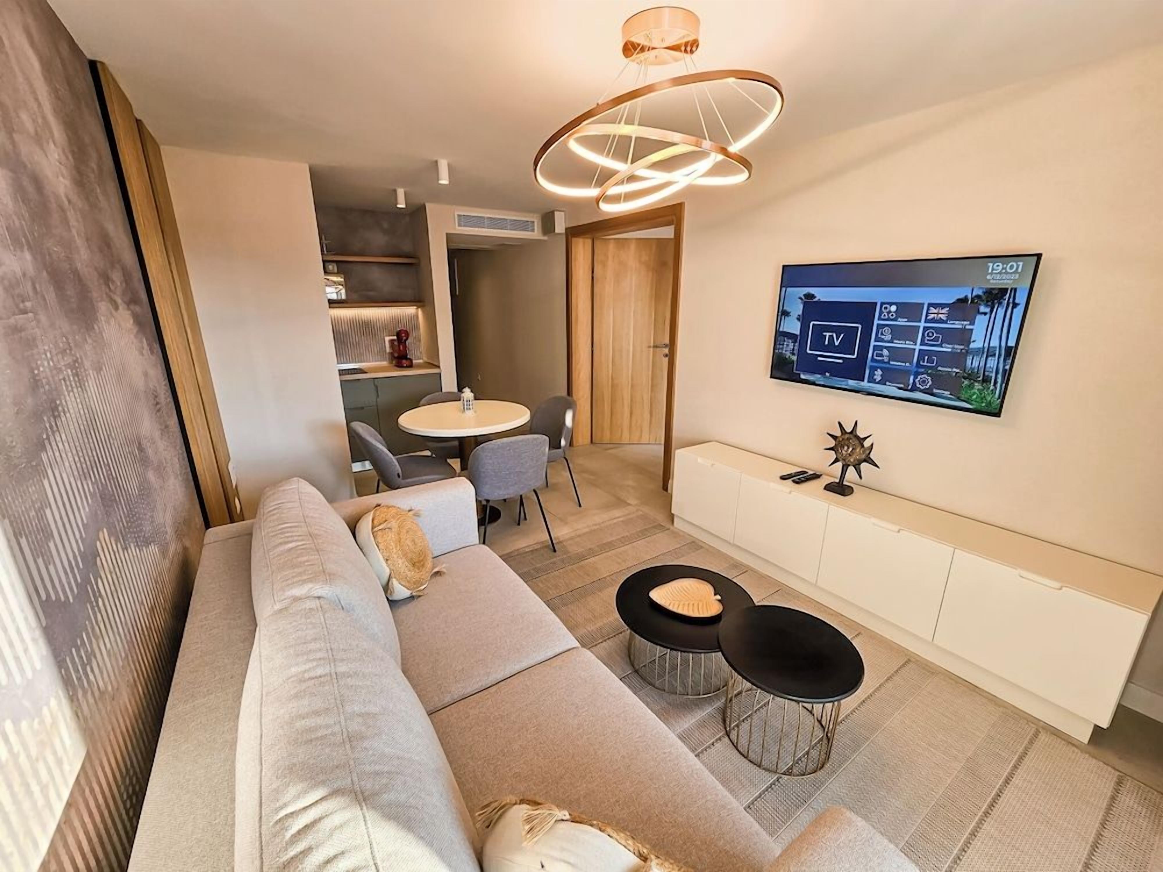 Ein modernes Wohnzimmer mit einer Couch, zwei runden schwarzen Tischen, einem an der Wand befestigten Fernseher und einer Holztür, die in einen anderen Raum führt.
