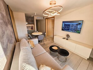 A modern living room with a sofa, two black round tables, a TV mounted on the wall, and a wooden door leading to another room. - The New Mediteran Hotel Villa Edition (Ulcinj)