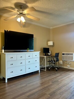 Ein Zimmer mit Deckenventilator, einem Flachbildfernseher auf einer weißen Kommode, einem Schreibtisch mit Computer und einer Klimaanlage.