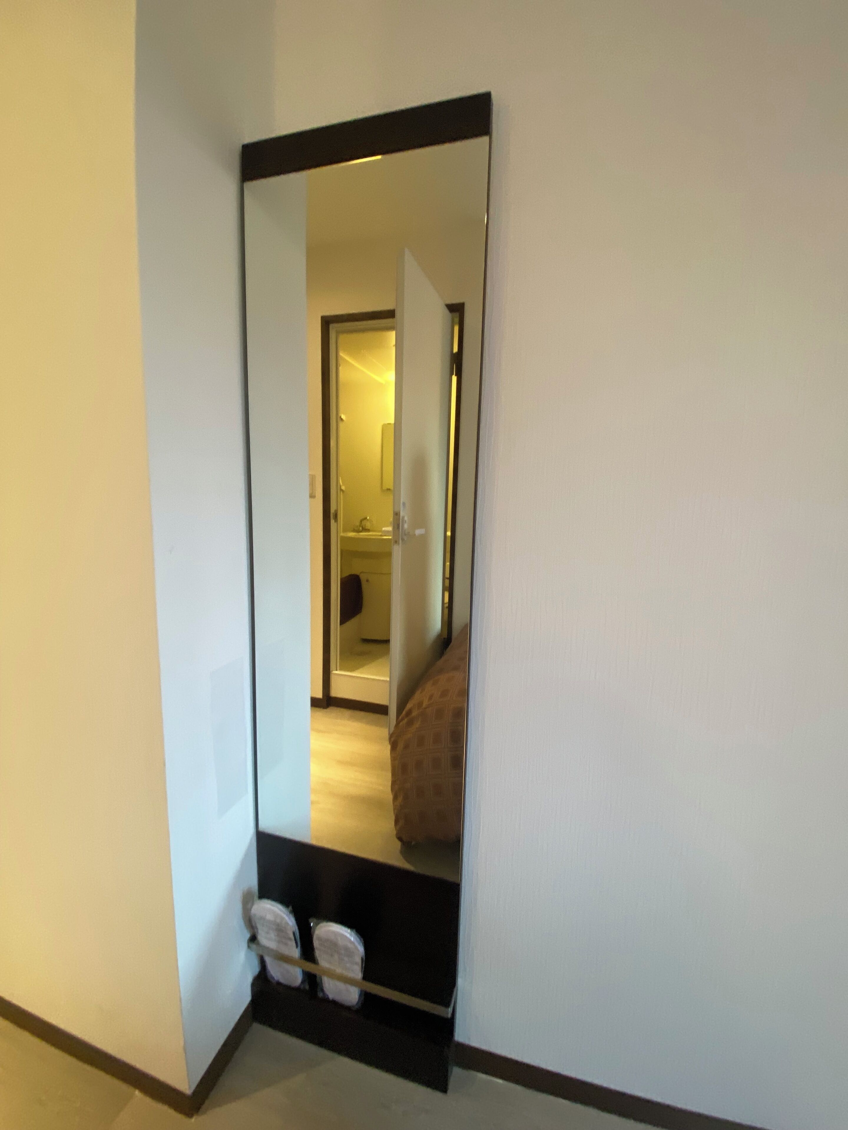 A hallway with a large mirror reflecting a room with a bed and a door leading to another room.