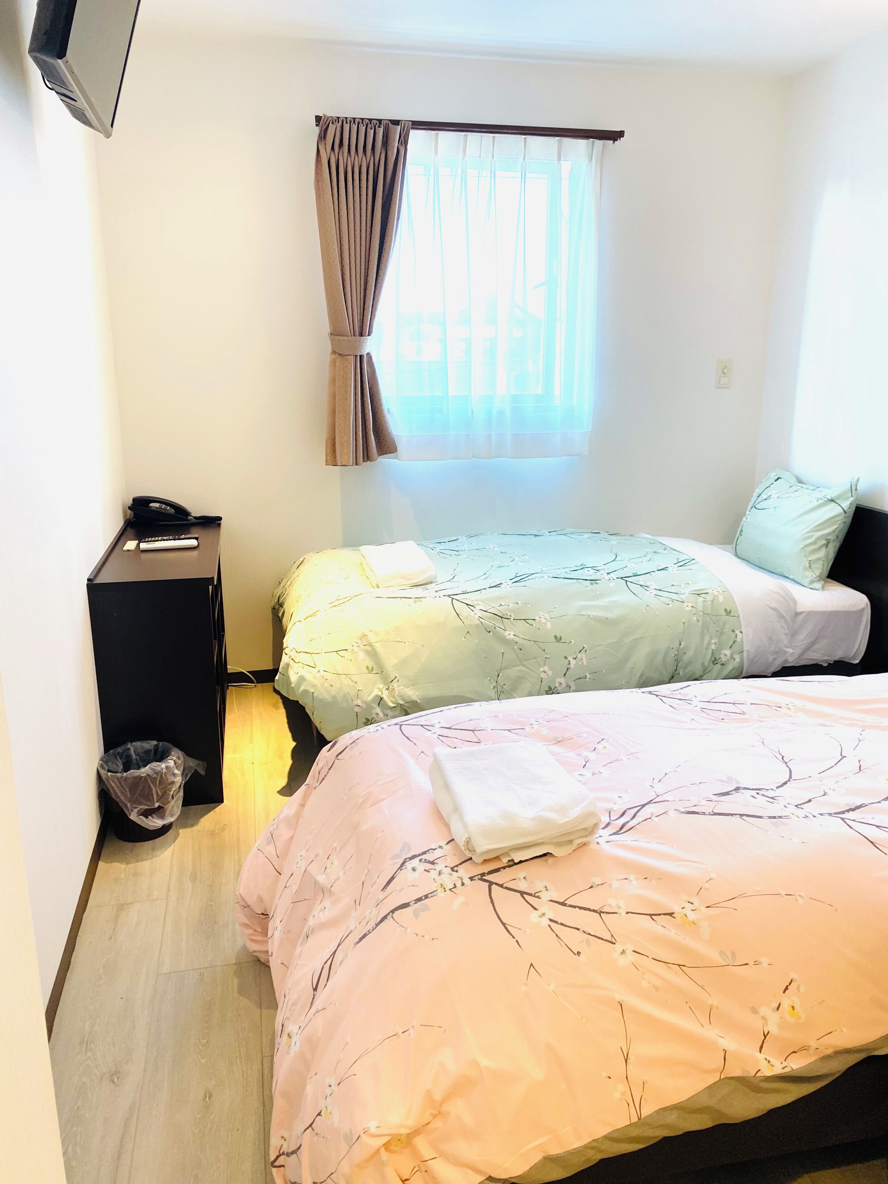 A bedroom with two beds, a bedside table with a phone, a trash bin, and a window with curtains.