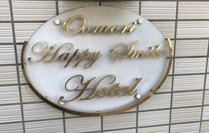 Um sinal circular com as palavras 'Omori Happy Smile Hotel' numa fonte cursiva, fixado numa parede com textura.