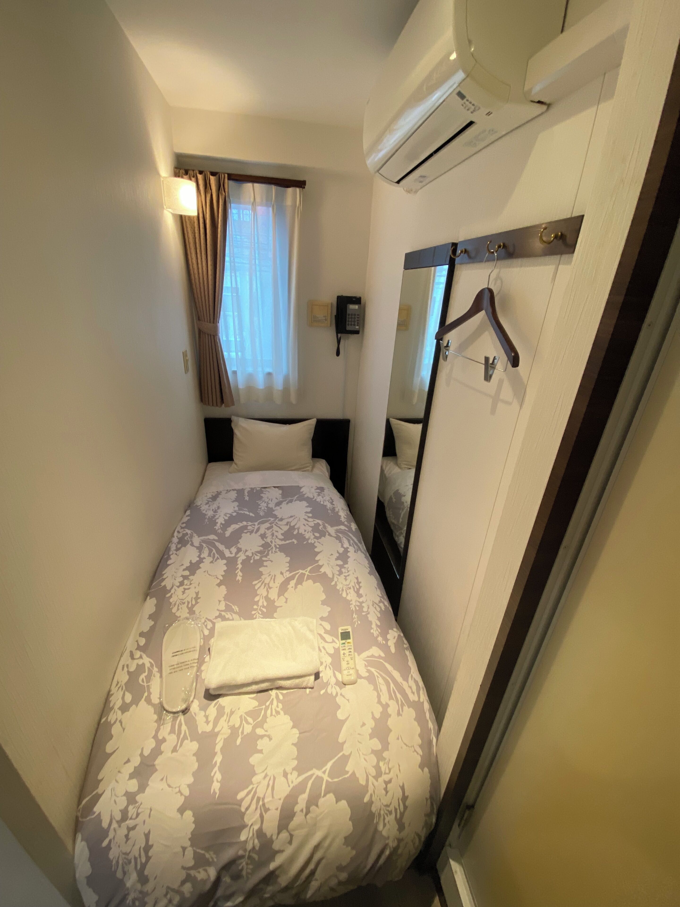 Une petite chambre d’hôtel avec un lit simple, un téléphone sur la table de chevet, un miroir et un porte-manteau.