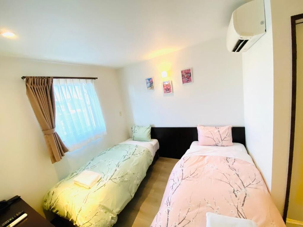 Ein Zimmer mit zwei Betten, einem mit Vorhängen versehenen Fenster, einer Wand-Klimaanlage und einem kleinen Tisch mit Lampe.