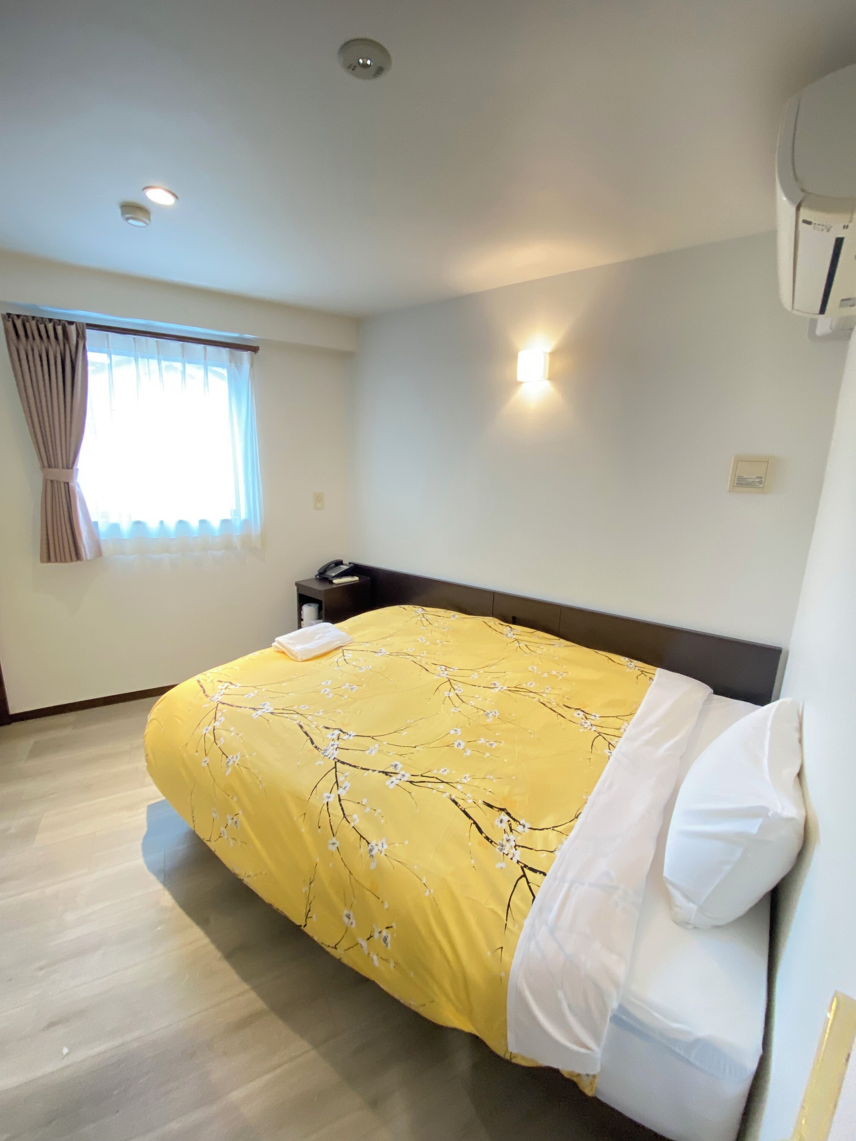 A hotel room with a bed, a nightstand, a window with curtains, a wall-mounted air conditioner, and a ceiling light.