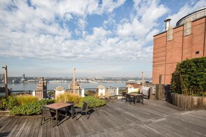 A rooftop patio with a view of a river and cityscape, featuring a wooden deck, outdoor seating, and a brick building. - Blueground | Upper West Side, rooftop, nr Central Park (New York)