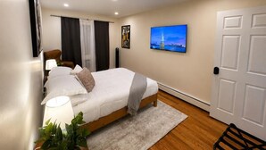 Ein Schlafzimmer mit Bett, Nachttischlampe, Vorhängen, einem an der Wand montierten Fernseher und einer Tür.