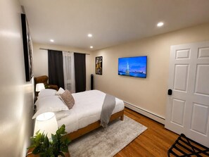 Ein Schlafzimmer mit Bett, Nachttischlampe, Vorhängen, einem an der Wand montierten Fernseher und einer Tür.