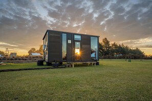 Una cabaña moderna y compacta con puerta de cristal, ubicada en un campo cubierto de césped con un atardecer de fondo.