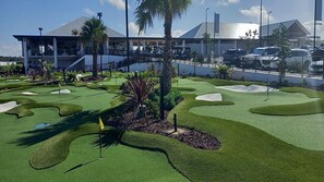 Ein Golfplatz mit einem modernen Gebäude im Hintergrund, Palmen und blauem Himmel.