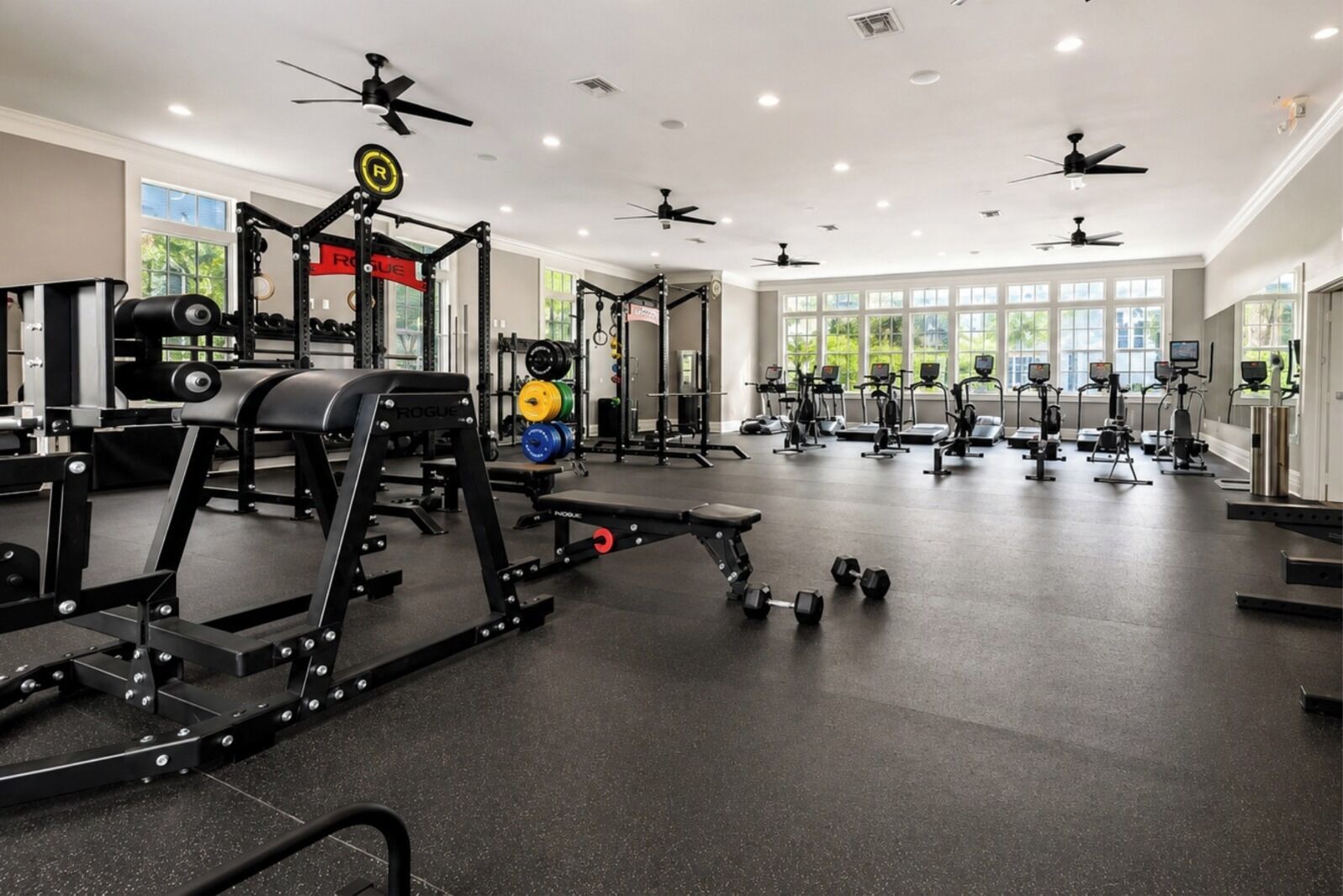 Une salle de sport équipée de divers appareils d’exercice, de ventilateurs de plafond et de grandes fenêtres.