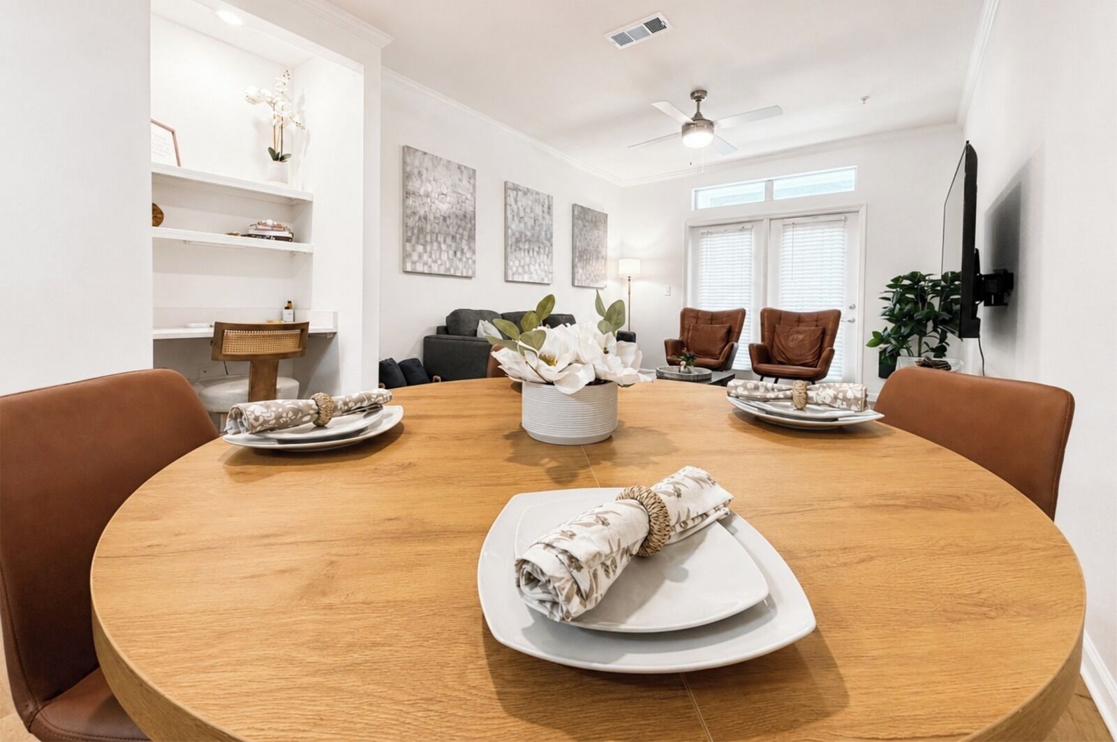 A dining room with a round wooden table, brown leather chairs, white plates, and a centerpiece with white flowers.