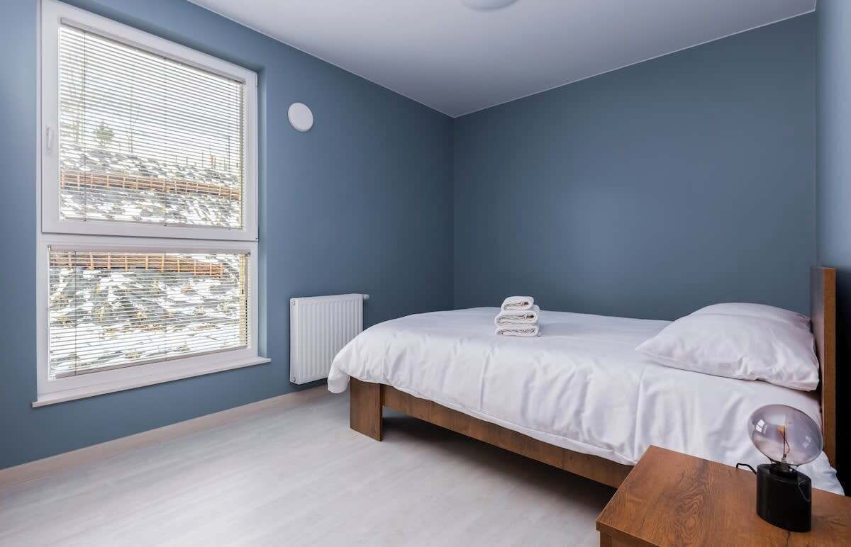 A bedroom with a bed, a bedside table, a radiator, and a window with blinds.